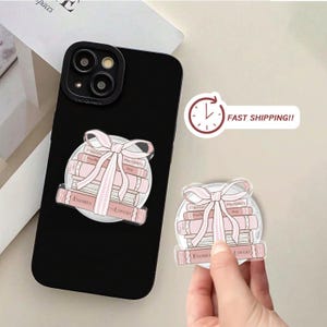 May include: Black phone case with a pink and white sticker showing a stack of books tied with a bow. The books are labelled "Enemies to Lovers" and "Forged in Proximity".