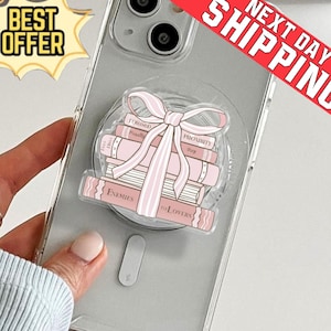 May include: A clear phone case with a pop-up grip featuring a stack of pink books tied with a pink bow. The books have titles like "Enemies to Lovers" and "Proximity". The phone case is on a light background.