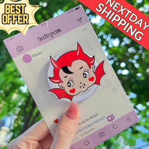 May include: A clear phone grip featuring a cartoon devil with red horns, wings, and heart-shaped cheeks. The grip is attached to a card designed to look like an Instagram post. The card has the text "Instagram" and "Album."