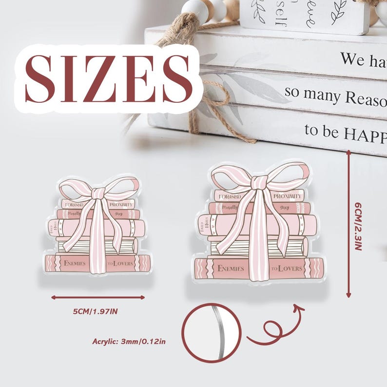 May include: Two acrylic stickers shaped like stacks of books with a pink bow. The stickers are 5 cm and 6 cm tall. The text "ENEMIES to LOVERS" is visible on the bottom book. The word "SIZES" is in the upper left. Acrylic is 3mm thick.