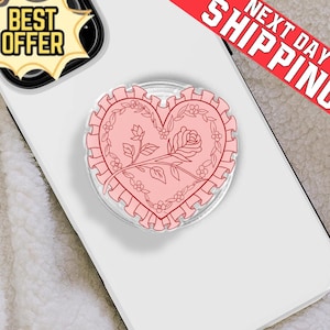 May include: A pink heart-shaped phone grip with a floral design, including a rose and small flowers, is centered on a white phone. The grip has a ruffled edge. The words "BEST OFFER" are in a yellow and black banner.