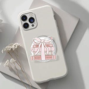 May include: A white phone case featuring a design of pink books and a bow. The books have titles including "Enemies to Lovers". The phone case is on a light grey surface.