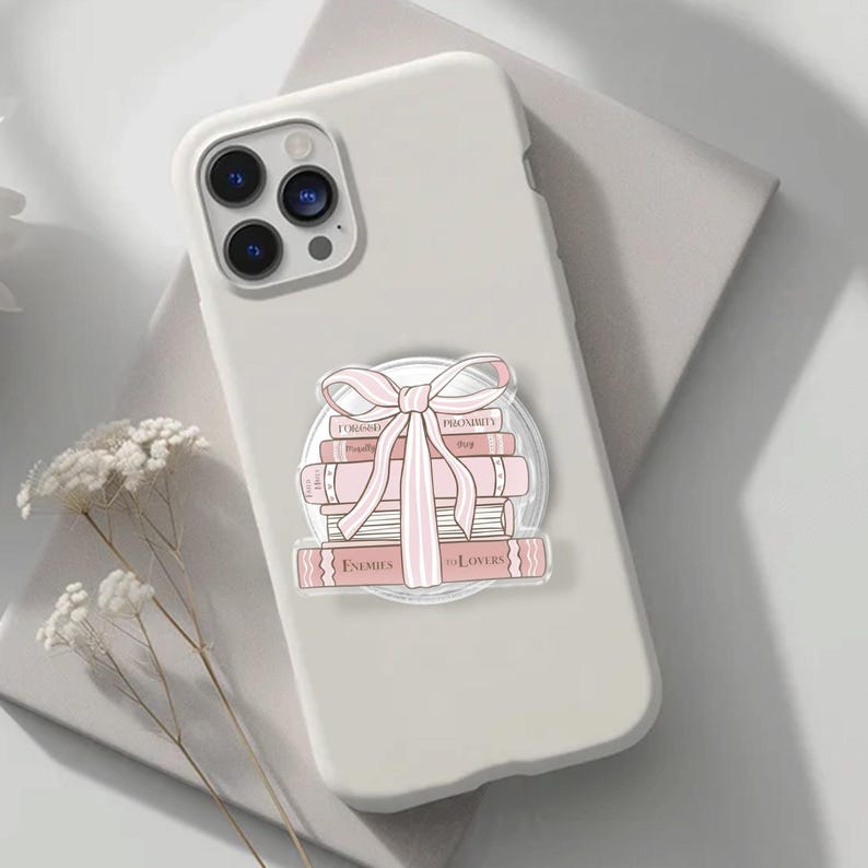 May include: A white phone case featuring a design of pink books and a bow. The books have titles including "Enemies to Lovers". The phone case is on a light grey surface.
