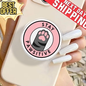 Può includere: Un supporto per telefono rotondo con un design rosa e bianco. Il centro presenta una zampa di gatto grigia con cuscinetti rosa e le parole "STAY PAWSITIVE". Il supporto è attaccato a un telefono beige.