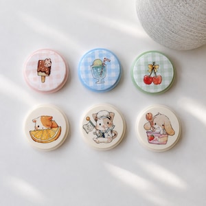 May include: Six round, decorative magnets with cute illustrations. Designs include a bunny on a popsicle, a sundae, cherries, a cat on an orange slice, a cat with a flag, and a bunny with strawberries. Each magnet has a different pastel background.
