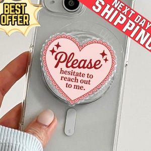Könnte beinhalten: Transparente Handyhülle mit einem herzförmigen Pop-Socket in Rosa. Der Pop-Socket hat das Wort "Please" in Schreibschrift und den Satz "hesitate to reach out to me". Der Pop-Socket hat einen gewellten Rand und vier Sternakzente.