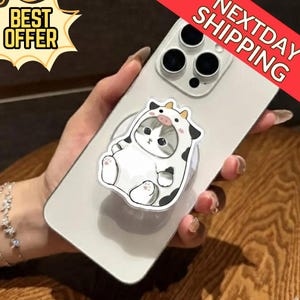 May include: A white smartphone with a pop-up grip featuring a cartoon cat in a cow costume. The grip is white with a clear border. The phone has a triple-lens camera. The image also includes the text "BEST OFFER" and "NEXTDAY SHIPPING".