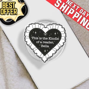 May include: A clear phone grip featuring a black heart design with a white frilly border. The heart has the text "This is the Kindle of a reader, Bella". A "Best Offer" sticker is in the top left.