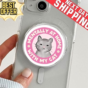 May include: A clear phone case with a pop socket featuring a gray cat illustration and the text "MENTALLY AT HOME WITH MY CAT" in a pink circle. The phone is held by a person.