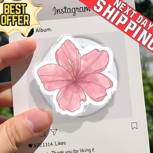 May include: A pink floral pop-up phone grip with a watercolor design. The grip is round and features a light pink flower with a white background. The image is on a white card with Instagram-themed graphics and the text "BEST OFFER" and "NEXT DAY SHIPPING".