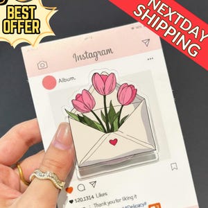 May include: A pink and white Instagram-themed sticker with a drawing of pink tulips in an envelope. The sticker has the text "Instagram" and "Album." The sticker is held by a hand. The sticker also has the text "BEST OFFER" and "NEXTDAY SHIPPING."