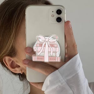 May include: A clear phone grip with a stack of pink books and a pink bow design. The books have text that reads "Forged", "Proximity", "Enemies", and "To Lovers". The phone is white.