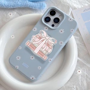 May include: A pale blue phone case decorated with white daisy motifs. A clear pop-socket features a stack of pink books tied with a pink ribbon. The books have the words "Enemies", "Lovers", "Forged", and "Proximity" printed on them.
