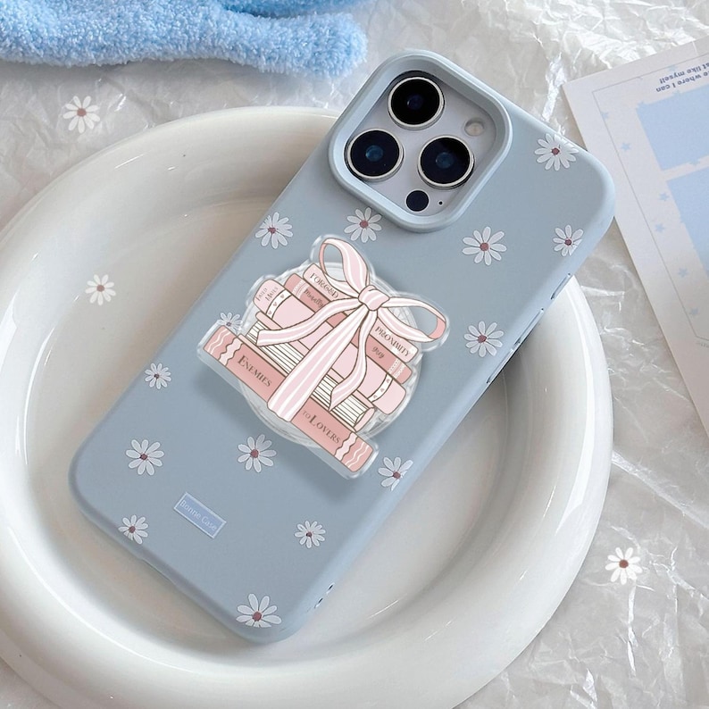 May include: A pale blue phone case decorated with white daisy motifs. A clear pop-socket features a stack of pink books tied with a pink ribbon. The books have the words "Enemies", "Lovers", "Forged", and "Proximity" printed on them.