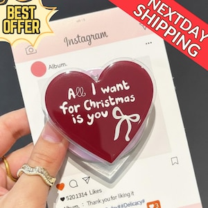 May include: A red heart-shaped phone grip with the text "All I want for Christmas is you" in white. The grip has a white bow graphic. The background is a social media-themed design.