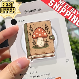 Könnte beinhalten: Ein Aufkleber mit einer Illustration eines Pilzes auf einem Buch. Das Buch ist braun mit grünen Akzenten und Blattmotiven. Der Aufkleber befindet sich auf einer weißen Karte im Instagram-Stil mit dem Text "Album" und "Instagram".