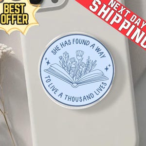 Puede incluir: Un soporte redondo para teléfono de color azul claro con el texto "SHE HAS FOUND A WAY TO LIVE A THOUSAND LIVES" que rodea un diseño floral y un libro abierto. El soporte está sobre una funda de teléfono gris claro.