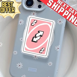 Op de afbeelding: Een lichtblauwe telefoonhoes met een madeliefjespatroon en een popsocket met een roze Uno reverse card design. De telefoonhoes heeft de tekst "Bonnie Case" onderaan. De afbeelding bevat ook een banner "Beste aanbieding" en "Volgende dag verzending".