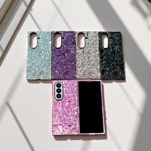 May include: A collection of iridescent phone cases in various colors, including light blue, purple, silver, and black. A pink phone case is open, displaying a black screen. The cases have a textured, shimmering surface.