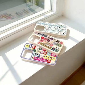 Puede incluir: Tres fundas de teléfono coloridas con diseños de teclado. La funda superior es azul claro con teclas blancas, la funda del medio es crema con teclas coloridas y la funda inferior es morada y rosa con teclas de personajes de dibujos animados.