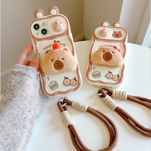 Puede incluir: Dos fundas de teléfono con un diseño de oso lindo. Las fundas son blancas con un borde rosa y presentan un oso de peluche 3D, un osito y ilustraciones de comida. También se ven correas de teléfono marrones y beige.