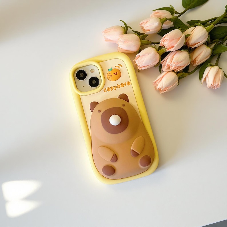 May include: A yellow phone case featuring a brown capybara design with a white button nose and small arms and legs. The case has the word "capybara" printed on it, along with a small orange graphic.