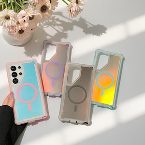 Puede incluir: Cuatro fundas de teléfono coloridas con un diseño de círculo magnético. Las fundas son en tonos de azul, naranja y gris, con un borde transparente. Una funda es sostenida por una persona. Un jarrón de flores está en el fondo.