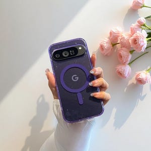 May include: A dark purple phone case with a clear, speckled design. The case has a circular purple accent with the letter "G" and a rectangular camera cutout. Pink roses are in the background.