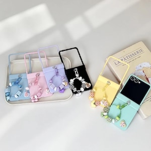 May include: A collection of colorful phone cases with attached straps and decorative charms. The cases are in various pastel colors, including blue, pink, purple, black, and yellow. Each case features a unique charm design, including bunny figures and checkered beads.