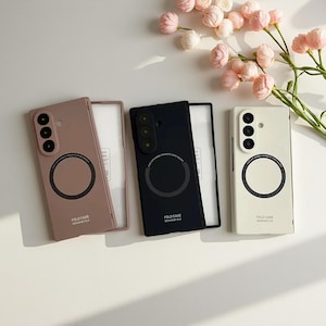 May include: Three phone cases in pink, black, and cream colors, each with a circular magnetic holder and a camera array. The cases are labeled "FOLD CASE" and "DESIGN BY ZLS". Pink flowers are in the background.