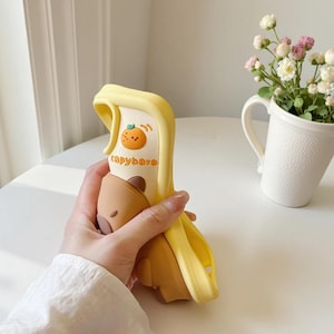 May include: A yellow and brown phone case featuring a capybara design. The case has a cartoon orange illustration and the word "capybara" printed on it. The case is held in a hand.