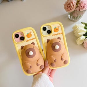 May include: Two yellow phone cases with a cartoon bear design. Each case features a brown bear with a white belly button, a small orange fruit, and the word "capybara". The cases are held by a person.