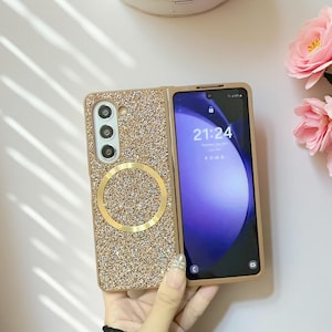 May include: A gold and glittery phone case with a gold ring and a phone screen displaying 21:24. The case is open, revealing the phone's screen. The case has a camera cutout with three lenses.