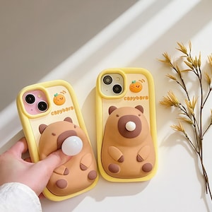 Puede incluir: Dos fundas amarillas para teléfono con un diseño de oso marrón. Las fundas tienen una cara de oso en 3D con una nariz blanca y pequeñas orejas y patas marrones. El texto "capybara" está impreso en la funda, junto con una pequeña ilustración de naranja.