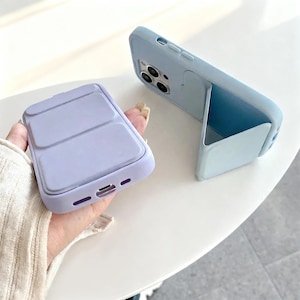 May include: Two pastel blue phone cases with a stand feature. One case is held in a hand, showing a magnetic charging pad. The other case is standing upright, showcasing the stand feature. The cases have a sleek, modern design.