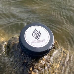 Può includere: Un piccolo barattolo rotondo nero con un'etichetta bianca con un disegno di pigna e la scritta "SHARE2PREPARE". Il barattolo è appoggiato su una roccia nell'acqua. L'acqua è leggermente increspata.