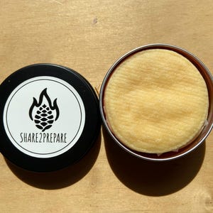 May include: A black tin with a white label featuring a flame and pinecone design, next to an open metal tin containing a round, yellow sponge. The text "SHARE2PREPARE" is printed on the label.