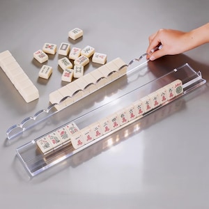 Puede incluir: Un juego de Mahjong con fichas de color marfil y símbolos rojos y negros. Las fichas están dispuestas en un soporte de acrílico transparente. Una mano ajusta el soporte. El juego está sobre una superficie plateada.
