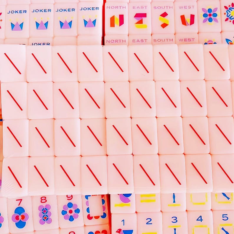 May include: A collection of pale pink mahjong tiles featuring various designs. Some tiles display the word JOKER with a crown icon, others show cardinal directions, and many feature a single red diagonal line or colorful floral and numeric patterns.