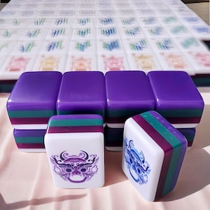 May include: A set of Mahjong tiles in purple and white. The tiles have a purple top and a striped edge in green, burgundy, and white. Some tiles feature a purple and white graphic of a mythical creature. More tiles are visible in the background.