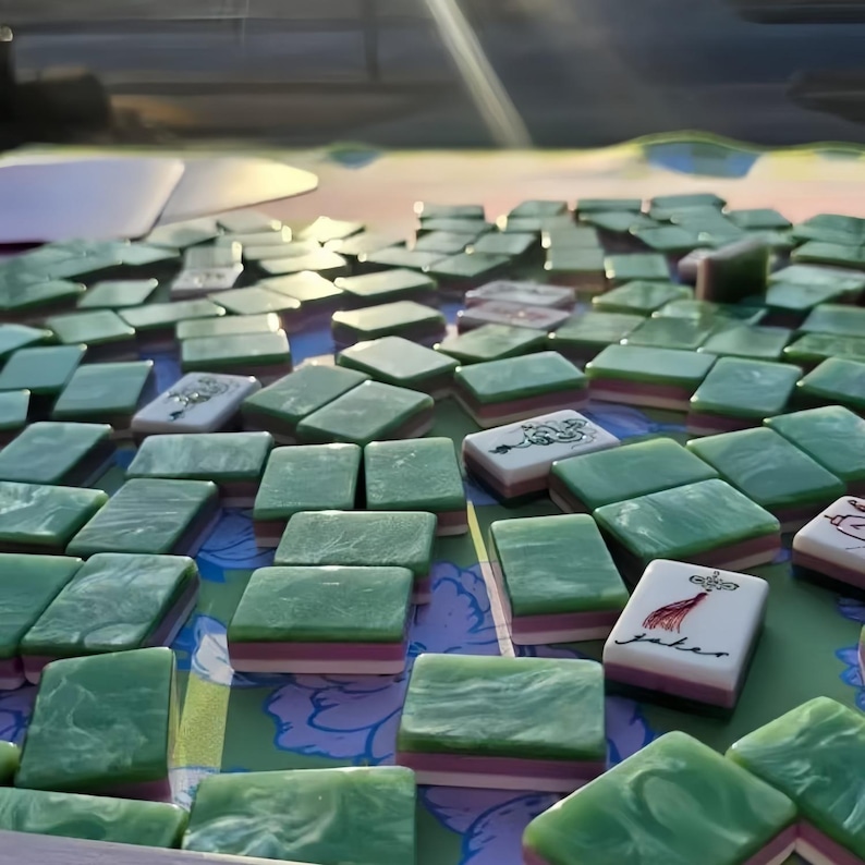 May include: A collection of green and white Mahjong tiles scattered on a floral patterned surface. The tiles have a marbled appearance and feature various symbols and characters. Some tiles have a pink and white stripe.