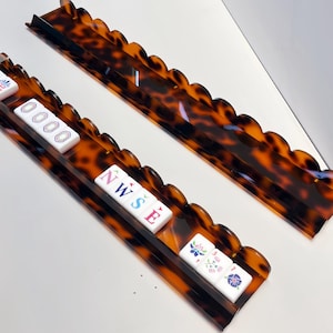 May include: Two long, rectangular mahjong tile racks with a scalloped top edge and a brown and black tortoiseshell pattern. One rack holds white tiles featuring colorful symbols, numbers, and the letters N, W, S, and E in red and blue ink.