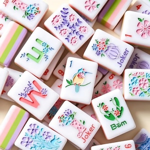 May include: A selection of white Mahjong tiles, each decorated with vibrant floral patterns and numbers. Some tiles show a bird, the word "Joker", or the number "1". The tiles are glossy and arranged in a random layout.