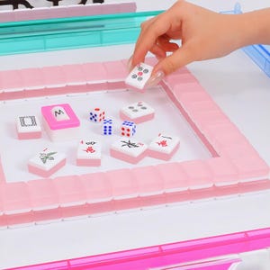 May include: A Mahjong set featuring translucent pink tiles with white faces, arranged in a square frame on a white surface. The set includes several loose tiles with black and red symbols, three small dice with blue and red dots, and a pink tile holder.