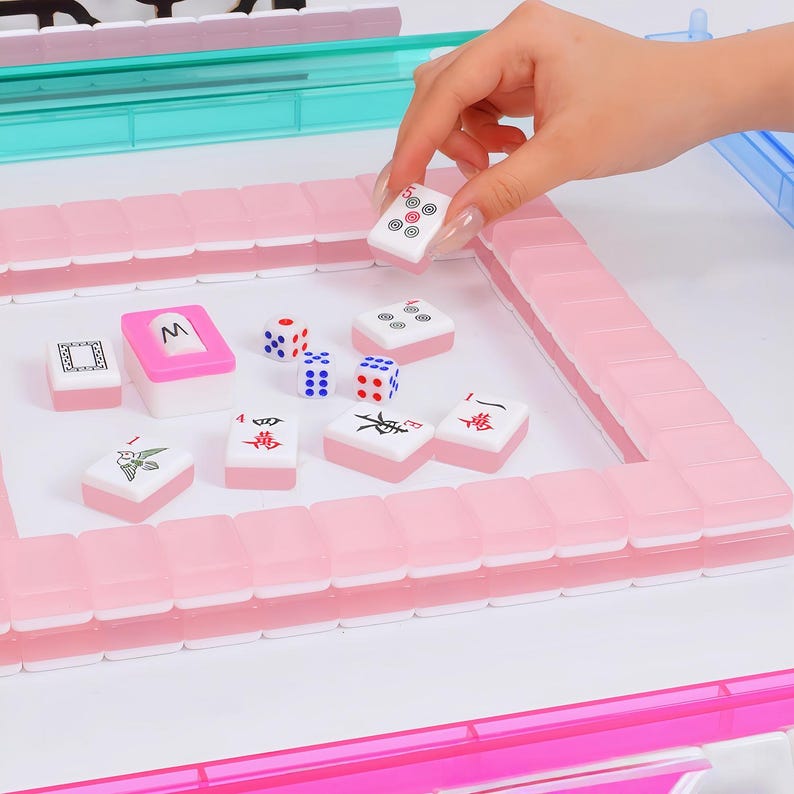 May include: A Mahjong set featuring translucent pink tiles with white faces, arranged in a square frame on a white surface. The set includes several loose tiles with black and red symbols, three small dice with blue and red dots, and a pink tile holder.