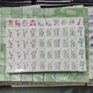 May include: A set of white Mahjong tiles with various illustrations and numbers, including birds, bamboo, and Chinese characters. The tiles are arranged on a green surface, with some tiles wrapped in plastic.