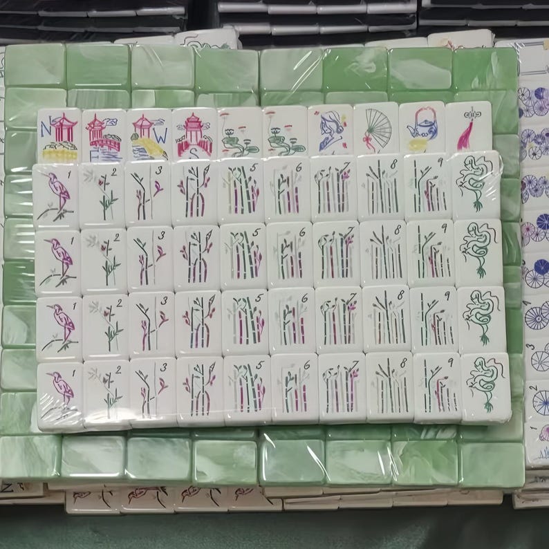 May include: A set of white Mahjong tiles with various illustrations and numbers, including birds, bamboo, and Chinese characters. The tiles are arranged on a green surface, with some tiles wrapped in plastic.