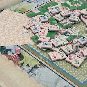 May include: A collection of Mahjong tiles with green and pink accents, scattered on a floral patterned game board. Two pink dice are visible, along with a decorative card. The game board has text that reads "PLAY RIGHT" and "FALLS LEFT".