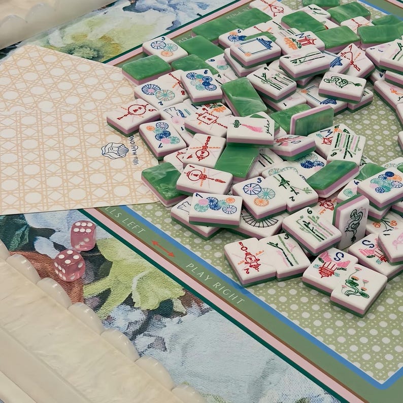 May include: A collection of Mahjong tiles with green and pink accents, scattered on a floral patterned game board. Two pink dice are visible, along with a decorative card. The game board has text that reads "PLAY RIGHT" and "FALLS LEFT".