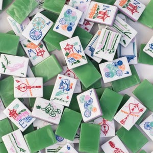 May include: A collection of white and green Mahjong tiles. The white tiles feature colorful illustrations of pagodas, flowers, and other symbols. The green tiles are solid and rectangular. The tiles are scattered, creating a visually appealing pattern.